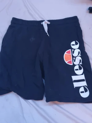 Svarta shorts från Ellesse med tryck - Svarta shorts från Ellesse med stort vitt logotryck och röd-orange detalj på ena benet. De har elastisk midja med vit snörning och är perfekta för en sportig look. Tillverkade i mjukt material som känns skönt mot huden.
