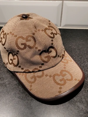 Beige keps med GG-mönster - Snygg beige keps från med det klassiska GG-monogrammet i brunt över hela kepsen. Skärmen har en mörkbrun kant i läderimitation och insidan är fodrad i ljus textil. Perfekt accessoar för dig som gillar exklusiv och trendig streetstyle. Väldigt fint skick!