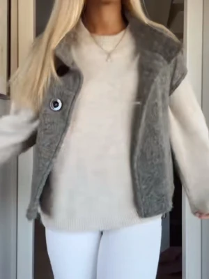 Grå stickad väst med stora knappar - Säljer en grå stickad väst med svarta knappar framtill. Västen har en bred krage och är ärmlös, perfekt att slänga över en tröja för en chill look. Materialet ser mjukt och mysigt ut, och färgen är lätt att matcha med olika outfits.