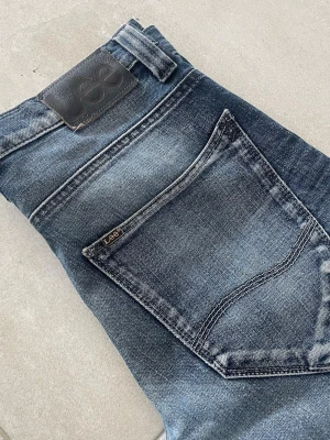 Lee blå jeans med tvättad look - Snygga blå jeans från Lee med klassisk femficksmodell och tydlig tvättad effekt. Jeansen har raka ben och normal midja, samt Lee-logga på bakfickan och läderpatch i midjan. Perfekta för en avslappnad och trendig stil. Mått 📏 Totallängd: 104cm Midjelängd: 40cm Innersömmlängd: 77cm Lårviddlängd: 25cm Skador 🔧: Tyget är lite löst vid baksidan av grenen, visas på bild  Storlek 🧍: W30/L34  Passform: Macky (Slim fit)  Jag kan förhandla mig till lägre pris.🙋💵 Om du har några frågor, vänligen skriv till