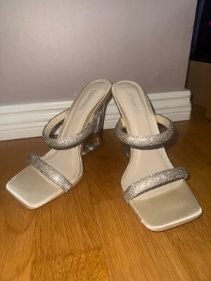 Beige pumps med glittriga band - Snygga pumps från NA-KD i beige med två glittriga remmar och transparent klack. Skorna har fyrkantig tå och en modern, stilren design som passar perfekt till trendiga outfits. Materialet är syntet med glittriga detaljer på banden.