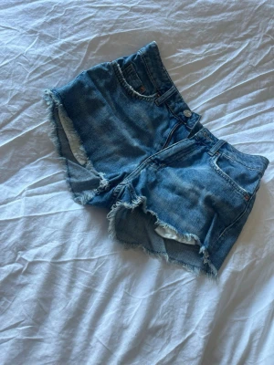Blå jeansshorts med fransig kant - Snygga blå jeansshorts med rå, fransig kant och klassisk femficksdesign. Shortsen har hög midja och är tillverkade i bomull, perfekta för varma sommardagar. Stängs med knapp och dragkedja framtill.