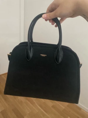 Svart handväska  - Stilren svart handväska med två rejäla handtag och guldfärgade detaljer. Väskan har en klassisk bågform och är tillverkad i mockaliknande material med detaljer i slätt konstläder. Perfekt för dig som gillar en tidlös och elegant look.