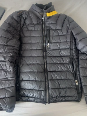 Svart dunjacka från Parajumpers - Snygg svart dunjacka från Parajumpers med gul detalj vid kragen och flera dragkedjeförsedda fickor. Jackan har quiltad design och hög krage som skyddar mot kyla. Perfekt för kalla vinterdagar och har en modern, sportig look.