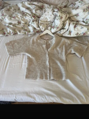 Beige kofta från Gina Tricot - Kort beige kofta från Gina Tricot med rund halsringning och knappar framtill. Koftan har ribbade muddar och är tillverkad i ett mjukt, stickat material. Perfekt att slänga över en topp för en chill look.
