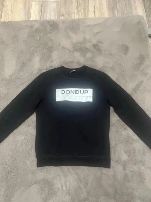 Svart sweatshirt från Dondup - Svart sweatshirt med rund hals och tryckt vit logga på bröstet från Dondup. Tröjan har långa ärmar och ribbade muddar vid ärmslut och nederkant. Perfekt för en avslappnad och stilren look.