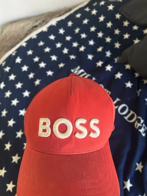 Röd keps från BOSS med brodyr - Säljer en röd keps från BOSS med vit broderad text framtill. Justerbar rem bak med randiga detaljer och metallspänne där BOSS-loggan syns. Klassisk böjd skärm och ventilerande hål upptill. Perfekt accessoar för en sportig och trendig look.