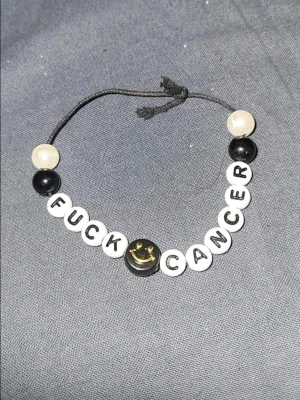 Fuck Cancer pärlarmband - Handgjort armband med vita och svarta plastpärlor på svart snöre. Bokstavspärlorna bildar texten 'FUCK CANCER' och i mitten finns en svart pärla med en gul smiley. Perfekt statement-accessoar för att visa stöd och styrka.