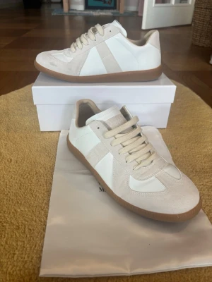 Maison Margiela vita gats - Snygga vita sneakers från Maison Margiela med beige mockadetaljer och klassisk gummisula. Skorna har en stilren design med vita läderpaneler, ton-i-ton snörning och låg profil. Perfekta för dig som gillar minimalistisk och trendig stil.