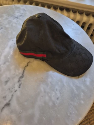 Svart keps från Gucci med röd/grön detalj - Snygg svart keps från Gucci med diskret GG-mönster i tyget och klassisk röd och grön rand på sidan. Kepsen har böjd skärm och justerbar passform. Perfekt accessoar för en trendig och exklusiv look.