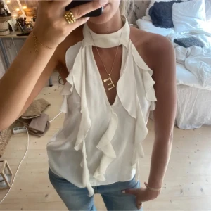 Vit volang topp med halterneck - Säljer en superfin vit halterneck topp med volangdetaljer framtill. Toppen är ärmlös och har en snygg v-ringning som ger en chic look. Perfekt att styla med jeans eller kjol för en trendig outfit.