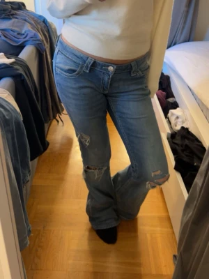 Blå bootcut jeans från Gina Tricot  - Säljer ett par blå bootcut jeans med snygga slitningar på båda benen. Jeansen har klassisk femficksdesign, låg midja och är tillverkade i denim. Perfekta för dig som gillar en avslappnad och trendig stil.