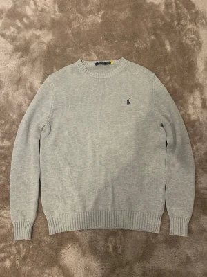 Ljusgrå stickad tröja från Polo Ralph Lauren - Ljusgrå stickad tröja från Polo Ralph Lauren med rund halsringning och diskret broderad logga i mörkblått på bröstet. Tröjan har ribbade muddar vid ärmslut och nederkant, vilket ger en klassisk och stilren look. Perfekt till jeans eller chinos.
