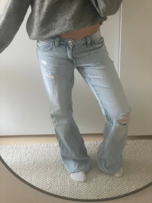 Ljusblå flare jeans - Säljer ett par ljusblå flare jeans med slitningar och hål på låret och knät. Jeansen har låg midja och klassisk femficksdesign. Perfekta för en avslappnad och trendig look. Aldrig använd!