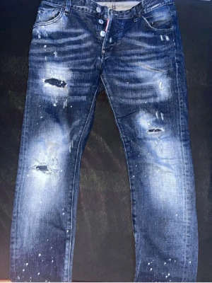 Mörkblå slitna jeans med knappar - Säljer ett par mörkblå jeans med slitningar och hål på benen samt vita färgstänk för en cool, trendig look. Jeansen har knappgylf och klassisk femficksmodell. Perfekta för dig som gillar streetstyle och en avslappnad vibe.