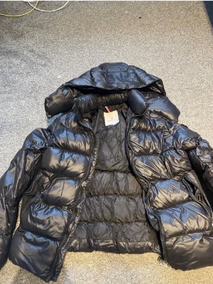 Svart pufferjacka med huva Moncler - Snygg svart pufferjacka från Moncler med glansig finish och stor vadderad huva. Jackan har breda kanaler, dragkedja framtill och två sidofickor med dragkedja. Perfekt för kalla dagar och riktigt trendig look.