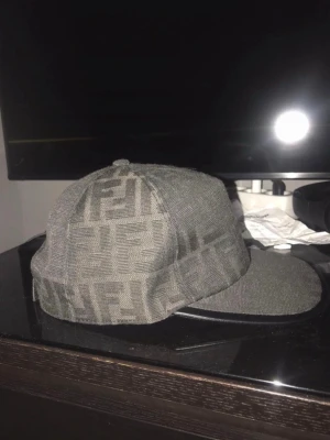 Grå Fendi keps med FF-mönster - Säljer en grå keps från Fendi med det klassiska FF-logomönstret över hela kepsen. Kepsen har böjd skärm och är tillverkad i ett tygmaterial som ger en exklusiv känsla. Perfekt accessoar för dig som gillar streetwear och designerplagg.