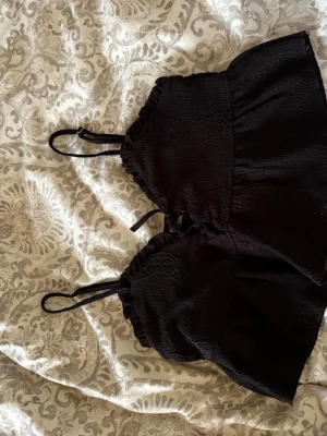 Svart spetsdetaljerat linne - En svart topp med tunna justerbara axelband och snygga spetsdetaljer. Toppen har en rynkad struktur och knytning framtill som ger en cool och trendig look. Perfekt för att matcha med jeans eller kjol.