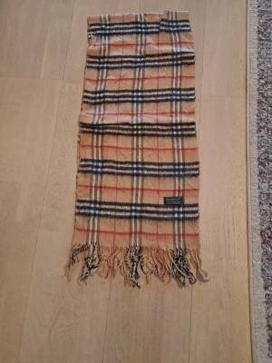 Beige rutig halsduk från Burberry - Klassisk beige halsduk från Burberry med ikoniskt rutmönster i svart, vitt och rött. Helt ny för endast 499 kr. Billugt pris. Halsduken har fransar i båda ändar och är tillverkad i 100% kashmir och ull. Perfekt accessoar för att lyfta din outfit med en tidlös och elegant touch. Tveka inte att kontakta om du är intresserad eller har några frågor! Först till kvarn som gäller!