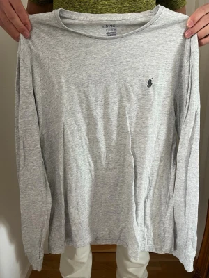 Grå långärmad tröja Polo Ralph Lauren - Säljer en stilren grå långärmad tröja från Polo Ralph Lauren. Tröjan har en klassisk passform, rund halsringning och den ikoniska lilla logotypen broderad på bröstet. Tillverkad i mjukt bomullsmaterial som känns skönt mot huden. Den är knappt använd och är i ett bra skick.