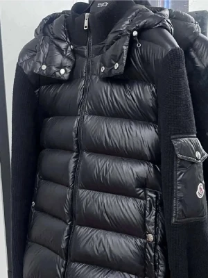 Svart pufferjacka från Moncler - Snygg svart pufferjacka från Moncler med glansig yta, stickade ärmar och huva med tryckknappar. Jackan har dragkedja framtill, fickor med knapp och Moncler-logga på ärmen. Perfekt för kalla vinterdagar och riktigt trendig look.