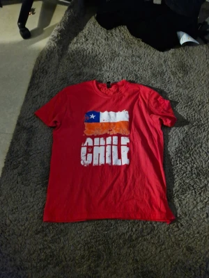Röd t-shirt med Chile-tryck - Säljer en röd t-shirt med tryck av Chiles flagga och texten 'CHILE' i vitt framtill. T-shirten har rund hals och korta ärmar, perfekt för dig som vill visa ditt stöd för Chile eller gillar coola tryck.