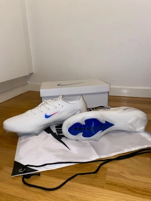 Nike vita fotbollsskor med blå swoosh - Snygga vita fotbollsskor från Nike med blå och rosa swoosh på sidan. Skorna har en modern, strömlinjeformad design och ovandel i syntetmaterial för bra bollkontroll. Sulan har blå detaljer och är anpassad för spel på gräs. Perfekta för dig som vill sticka ut på planen.