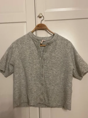 Ljusgrå stickad kortärmad kofta XXS - Supermjuk ljusgrå stickad kofta från H&M med korta ärmar och rund hals. Koftan har knäppning framtill med matchande knappar och ribbade kanter. Perfekt att slänga över en topp för en chill look.