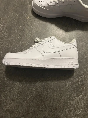 Nike Air Force 1 vita sneakers - Klassiska Nike Air Force 1 sneakers i helvitt läder med perforerad tå, vita skosnören och den ikoniska swoosh-loggan på sidan. Tjock sula med AIR-detalj och låg profil. Perfekta för en clean och stilren look. Helt nya.