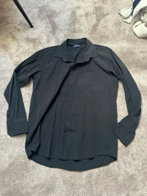 Svart klassisk skjorta med knappar - En stilren svart skjorta med lång ärm och knappar framtill. Skjortan har klassisk krage och rak passform, perfekt för en clean och enkel look. Tillverkad i mjukt bomullsmaterial som känns bekvämt mot huden. Enkel att matcha med olika outfits.