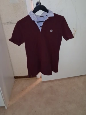 Vinröd kortärmad skjorta med mönstrad krage - Snygg vinröd skjorta med korta ärmar och vitmönstrad krage. Skjortan har en liten broderad logga på bröstet och klassisk knappslå. Perfekt för dig som vill ha en stilren men ändå unik look.