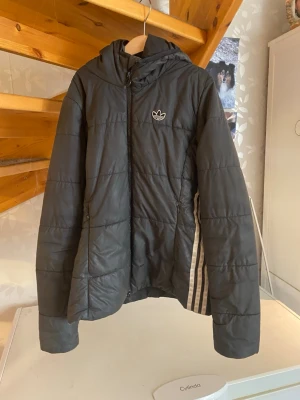 Svart dunjacka med huva från Adidas - Snygg svart dunjacka från Adidas med huva och dragkedja. Jackan har vita Adidas-ränder på sidan och den klassiska loggan på bröstet. Quiltad design och två sidofickor. Perfekt för kalla dagar och enkel att matcha med olika outfits.
