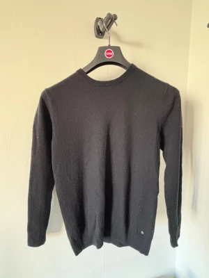 Svart stickad tröja i kashmir  - En enkel och stilren svart stickad tröja med rund halsringning och långa ärmar. Tröjan har ribbade muddar vid ärmslut och nederkant samt en liten metallbricka vid nederkanten. Perfekt basplagg till garderoben.