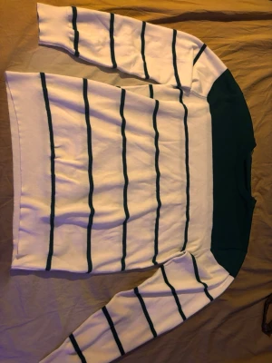 Vit stickad tröja med gröna ränder - Säljer en vit stickad tröja med tunna gröna horisontella ränder och en bred grön panel upptill vid axlar och hals. Tröjan har lång ärm och klassisk rund halsringning. Perfekt för dig som gillar enkel men snygg design.