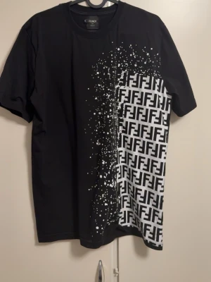 Svart och vit Fendi t-shirt - Snygg svart t-shirt från Fendi med korta ärmar och rund hals. Ena sidan har ett vitt grafiskt FF-mönster och stänk-effekt som ger en cool kontrast. Perfekt för dig som vill sticka ut med en unik designerstil.