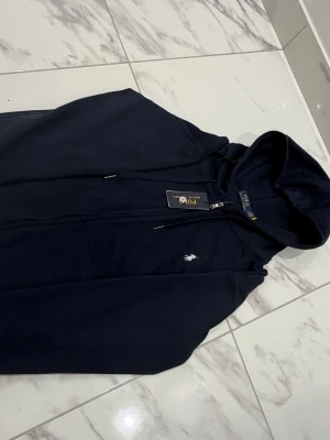 Marinblå hoodie från Polo Ralph Lauren - Snygg marinblå hoodie från Polo Ralph Lauren med dragkedja framtill och klassisk huva. Ikoniska vita loggan broderad på bröstet. Tillverkad i mjukt bomullsmaterial för skön känsla. Perfekt för en avslappnad och stilren look.