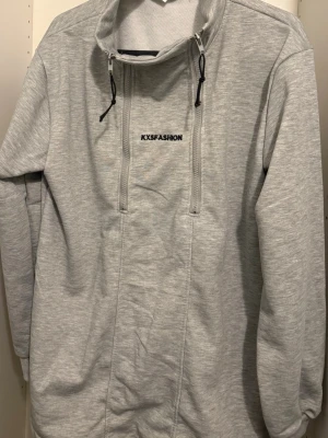 Ljusgrå hoodie från KXSFASHION - Ljusgrå hoodie från KXSFASHION med hög krage och dubbla dragkedjor framtill. Tillverkad i mjukt material och har en minimalistisk design med svart logotyp på bröstet. Perfekt för en avslappnad och stilren look.