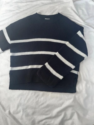 Svart stickad tröja med vita ränder - Snygg svart stickad tröja med breda vita horisontella ränder. Tröjan har rund halsringning och ribbstickade muddar. Perfekt för dig som gillar en enkel men trendig look.