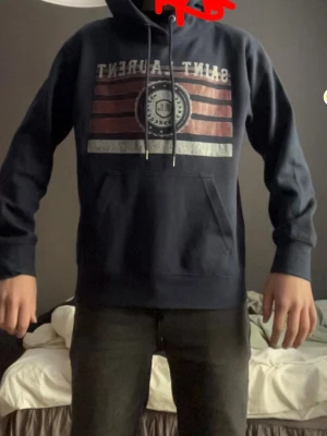 Mörkblå Saint Laurent hoodie - Snygg mörkblå hoodie från Saint Laurent med tryck framtill i rött, vitt och grått. Klassisk huva med dragsko och stor magficka. Perfekt för en avslappnad och cool stil. Storlek S