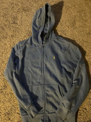 Blå zip hoodie från Polo Ralph Lauren - Snygg blå hoodie med dragkedja från Polo Ralph Lauren. Klassisk design med huva, fickor framtill och den ikoniska lilla loggan broderad på bröstet. Perfekt för en chill och avslappnad stil. Mjuk bomullskänsla och ribbade muddar vid ärmar och midja.