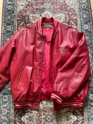 Röd läderjacka - Röd läder bomberjacka i varsity stil. Jackan har en snygg cropped oversize passform. Materialet är i kalvskinn. Storlek L, men passar mig bra som är 189cm och brukar använda storlek M på nästintill allt.