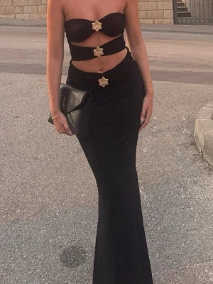 Svart långklänning med cut-outs - Trendig svart långklänning med cut-outs och dekorativa guldfärgade detaljer framtill. Klänningen är axelbandslös och har en figurnära passform som framhäver siluetten. Perfekt för dig som vill sticka ut med en unik och modern look.