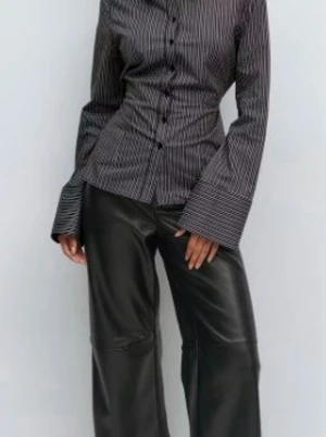 Randig svart skjorta med knytband - Snygg svart skjorta med vita ränder och knytband i midjan. Skjortan har långa ärmar, klassisk krage och knappar framtill. Tillverkad i ett mjukt bomullstyg som känns bekvämt och stilrent. Perfekt för dig som gillar en clean och modern look.