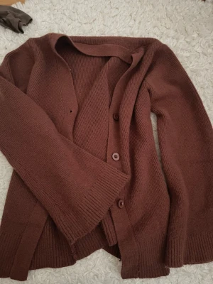 Brun stickad kofta med knappar - Mysig brun stickad kofta med långa ärmar och ribbade muddar. Koftan har en enkel design med knäppning framtill och mjukt material som känns skönt mot huden. Perfekt att slänga på sig när det är lite kyligt ute.