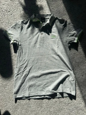 Grå pikétröja från BOSS - Snygg grå pikétröja från BOSS med klassisk krage och knappar framtill. Tröjan har diskreta gröna detaljer på krage och ärmslut samt BOSS-logga broderad på bröstet. Perfekt till jeans eller chinos för en clean look.