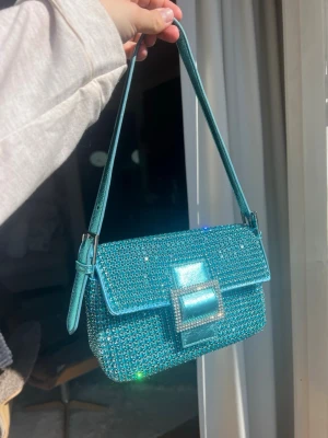Glittrig turkos handväska med spänne - Säljer en glittrig turkos handväska med rektangulär form och ett stort dekorativt spänne framtill. Väskan är täckt av små glittrande stenar och har ett justerbart axelband. Perfekt för dig som vill sticka ut med accessoarer.