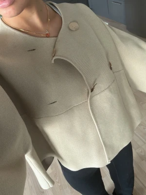 Beige cape-kappa från Zara - Stilren beige cape-kappa från Zara med oversized passform och guldiga knappar framtill. Kappan har en öppen, svepande design och är tillverkad i ett mjukt, tjockt tyg som ger en lyxig känsla. Perfekt för lager-på-lager och trendiga outfits. 