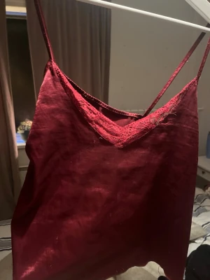 Vinrött linne med spetsdetaljer - Säljer ett vinrött pyjamaslinne med tunna axelband och spetsdetaljer vid urringningen. Linnet har en mjuk och glansig finish, perfekt för mysiga kvällar hemma. Materialet känns lent mot huden och ger en avslappnad känsla.