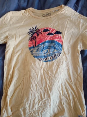 Gul t-shirt med tryck från Produkt - Säljer en gul t-shirt från Produkt i ekologisk bomull. Framsidan har ett färgglatt tryck med palmer, vågor och texten 'Good Vibes' i blått och rosa. Klassisk rund hals och korta ärmar. Perfekt för dig som gillar chill strandstil.