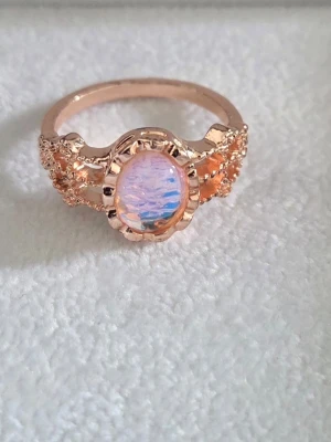Roséguldfärgad ring med opalsten - Aldrig använd. Unik ring i roséguld-look med en oval, skimrande opal-liknande sten i mitten. Ringen har detaljerade sidor med små pärlliknande dekorationer som ger en lyxig och trendig känsla. Perfekt för dig som vill sticka ut med accessoarer. Inre diameter: 16,5 (US 6) 